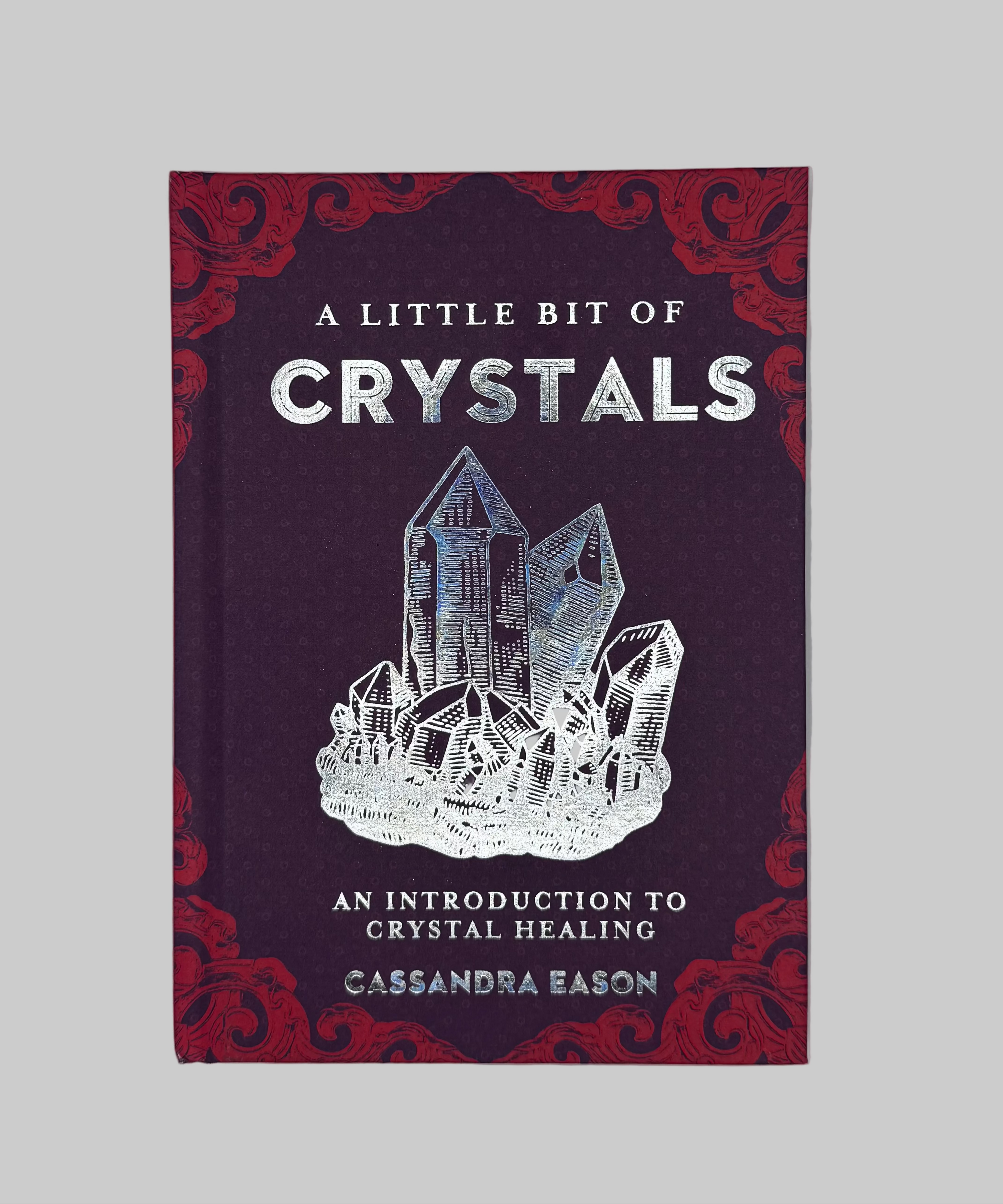 The book cover of 'A Little Bit of Crystals' by Cassandra Eason, featuring a deep purple background with red ornate corner patterns and a white crystal cluster illustration in the center.