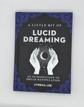 A book titled 'A Little Bit of Lucid Dreaming' by Cyrena Lee, featuring a black cover with blue ornate corner details and a white illustration of hands holding a crescent moon.