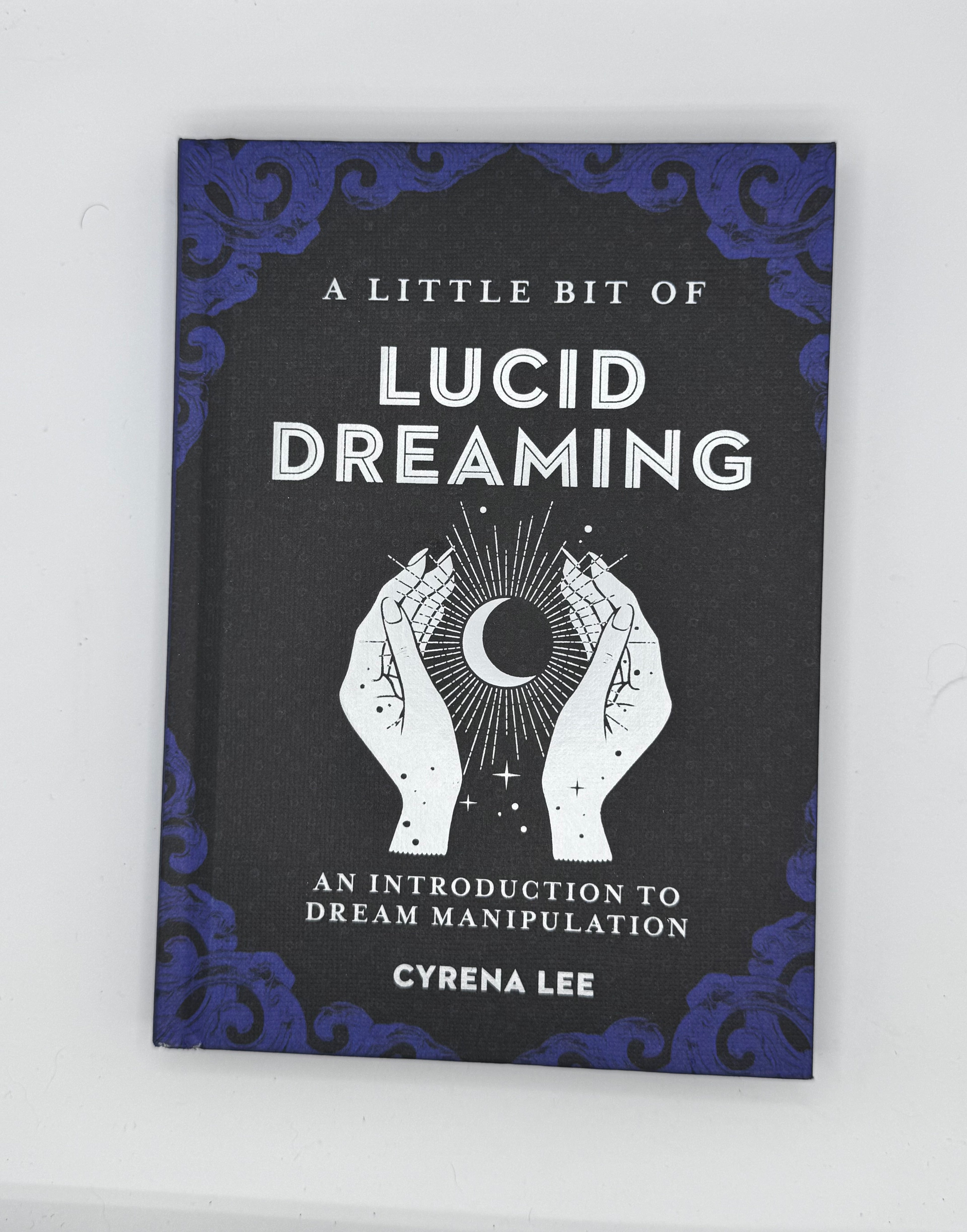 A book titled 'A Little Bit of Lucid Dreaming' by Cyrena Lee, featuring a black cover with blue ornate corner details and a white illustration of hands holding a crescent moon.