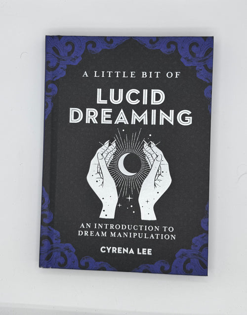 A book titled 'A Little Bit of Lucid Dreaming' by Cyrena Lee, featuring a black cover with blue ornate corner details and a white illustration of hands holding a crescent moon.