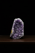 A raw amethyst geode specimen with dense purple crystal points and a small gold mineral inlay, standing upright on a black surface against a black background.